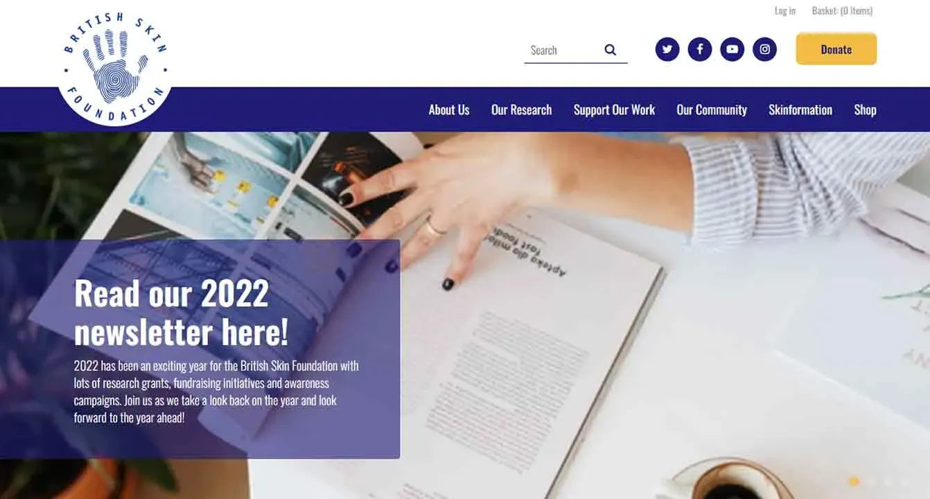 A person is pointing at a British Skin Foundation newsletter on a desk next to a pen and a magazine, with the foundation’s website header visible, featuring navigation links and a yellow "Donate" button.