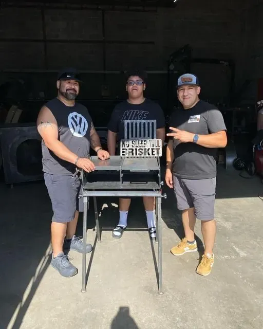 Tres personas están alrededor de una parrilla de metal en un garaje, con una persona usando pantalones cortos y una camiseta con un signo de paz, y otra persona en ropa deportiva, mientras que la parrilla tiene un letrero que dice "In Sleep Till Brisket."