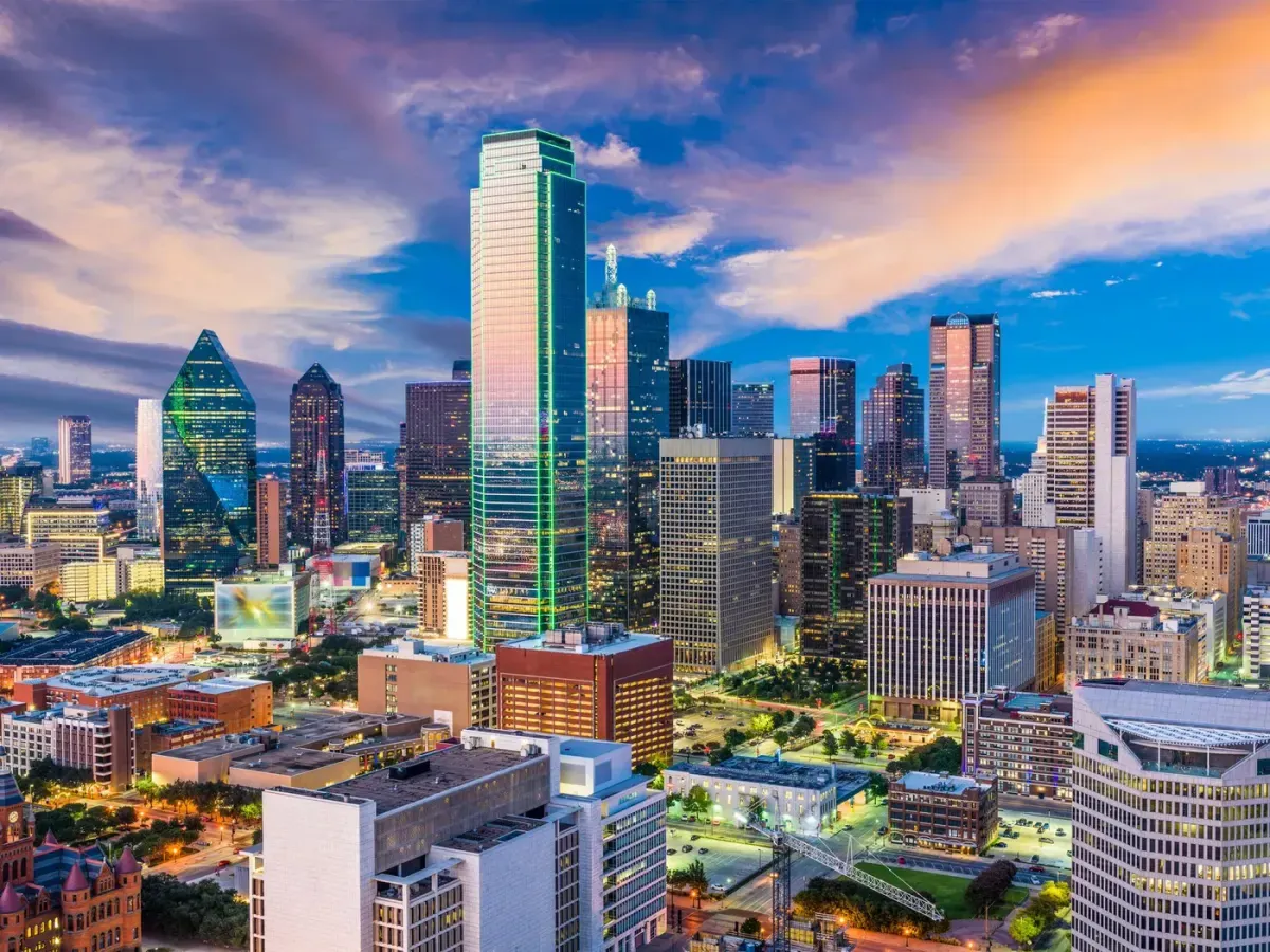Panorama de Dallas al atardecer, mostrando altos rascacielos con luces vibrantes que contrastan con un cielo colorido lleno de nubes naranjas y púrpuras.