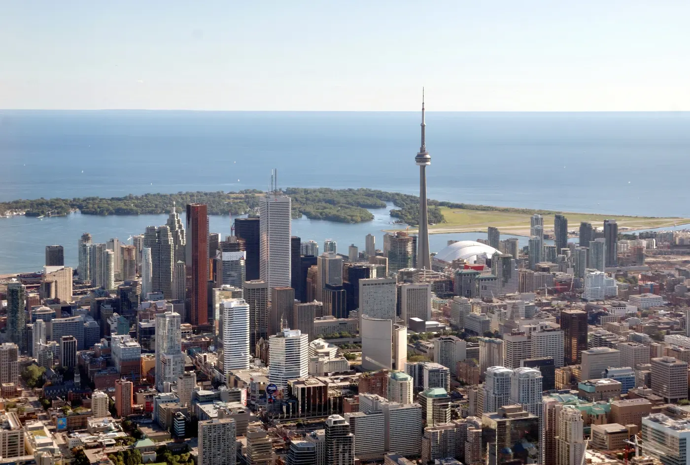 Vista aérea de Toronto, mostrando el horizonte con la icónica Torre CN, rodeada de modernos rascacielos y bordeada por las serenas aguas del Lago Ontario en un día despejado.