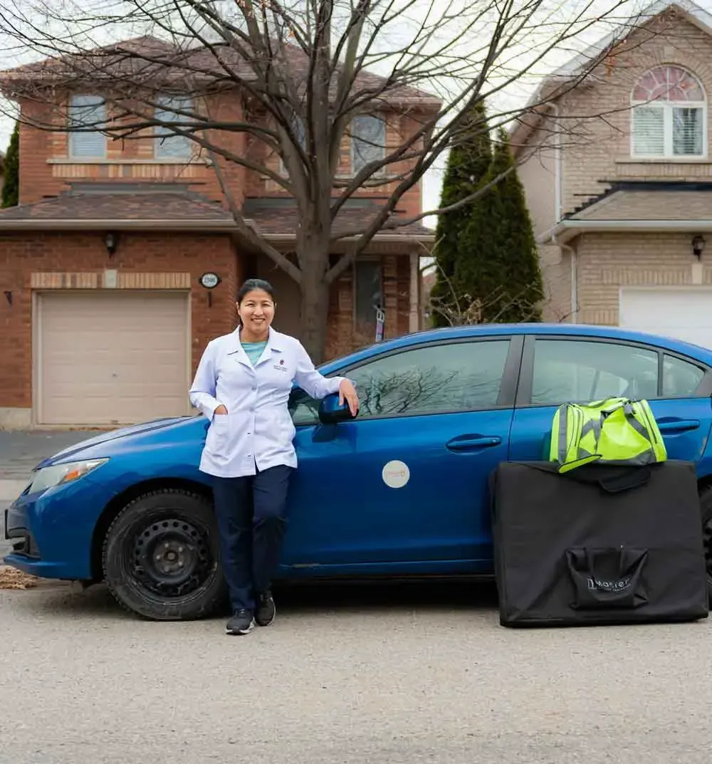 Una persona con una bata blanca de laboratorio y pantalones oscuros está de pie junto a un sedán azul en una calle residencial, con dos casas de ladrillo al fondo y un gran estuche negro y una bolsa verde neón colocados en el suelo cerca.