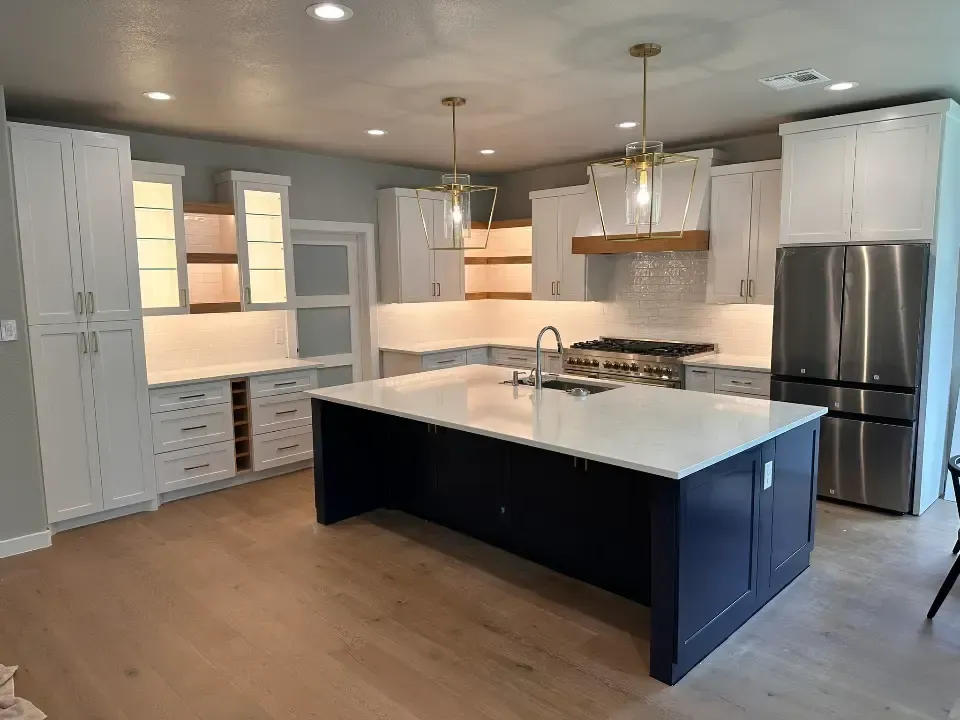 Una cocina moderna con una gran isla con encimera de cuarzo blanco, base azul oscuro y fregadero integrado, rodeada de elegantes gabinetes blancos, un refrigerador de acero inoxidable y luces colgantes, todo sobre un suelo de madera clara.