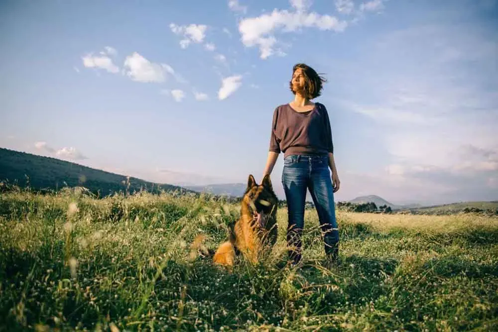 Mujer en jeans de pie junto a su perro en un prado iluminado por el sol con colinas distantes