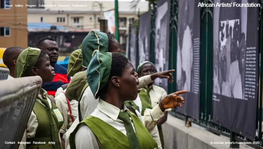 A group of students in green uniforms attentively observe and point at black-and-white photographs displayed outdoors, showcasing an educational or cultural event focused on history or art.