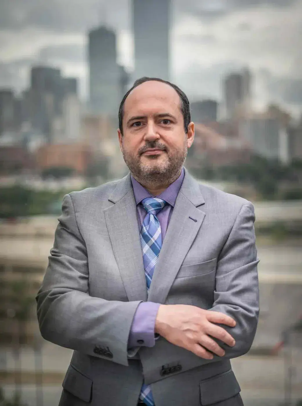 Un hombre en un traje gris con una camisa morada y una corbata a cuadros se para con confianza con los brazos cruzados contra un fondo de paisaje urbano desenfocado.