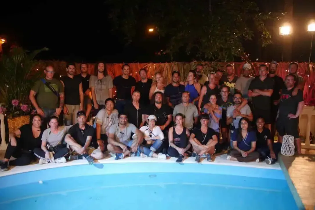 Un gran grupo de personas reunidas para una foto grupal junto a una piscina bien iluminada por la noche, rodeada de plantas tropicales y un ambiente cálido.