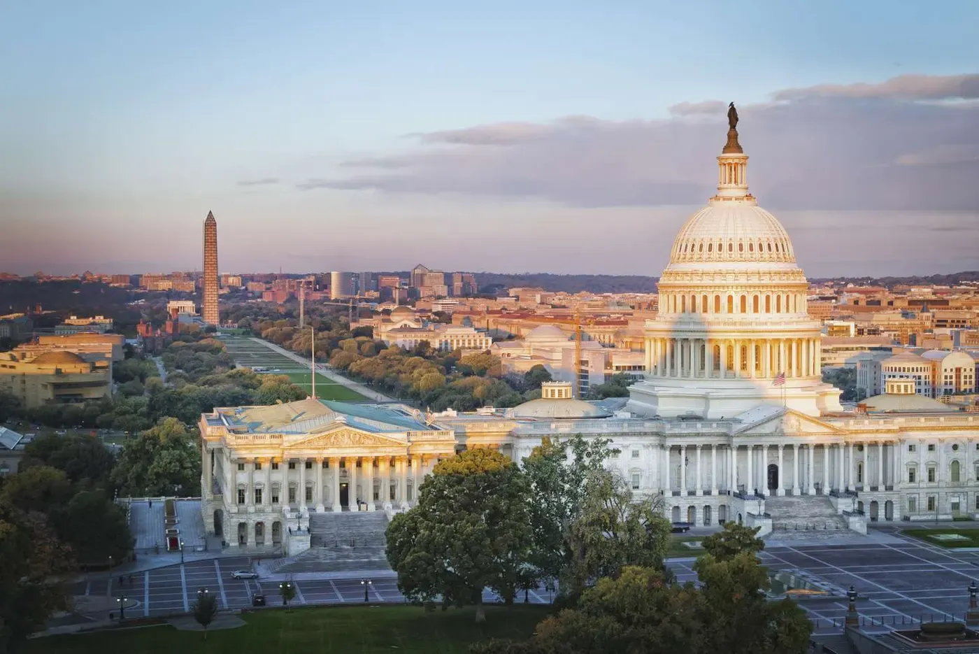 The image captures a panoramic view of the iconic United States Capitol Building at sunset, with the Washington Monument and National Mall stretching into the distance, framed by vibrant autumn foliage.