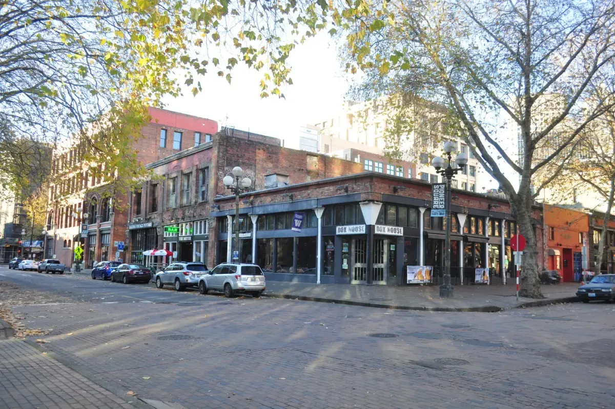 A scenic view of a historic urban street corner on a sunny day, featuring brick buildings with vintage architecture, parked cars along the cobblestone street, and tall trees with autumn leaves, embodying the charm of an old city neighborhood.