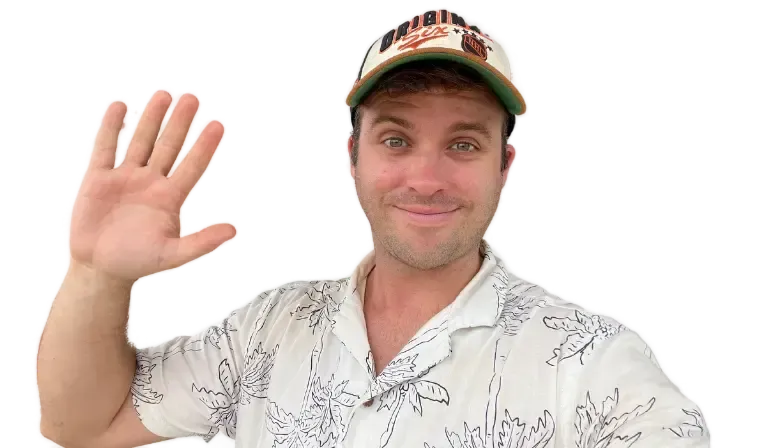 A smiling person in a hat waves with one hand, wearing a light-colored shirt against a neutral background.