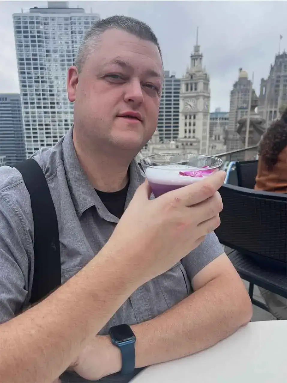 A person in a gray shirt enjoys a colorful cocktail on a rooftop bar with a city skyline view, including tall buildings and a historic clock tower, on an overcast day.