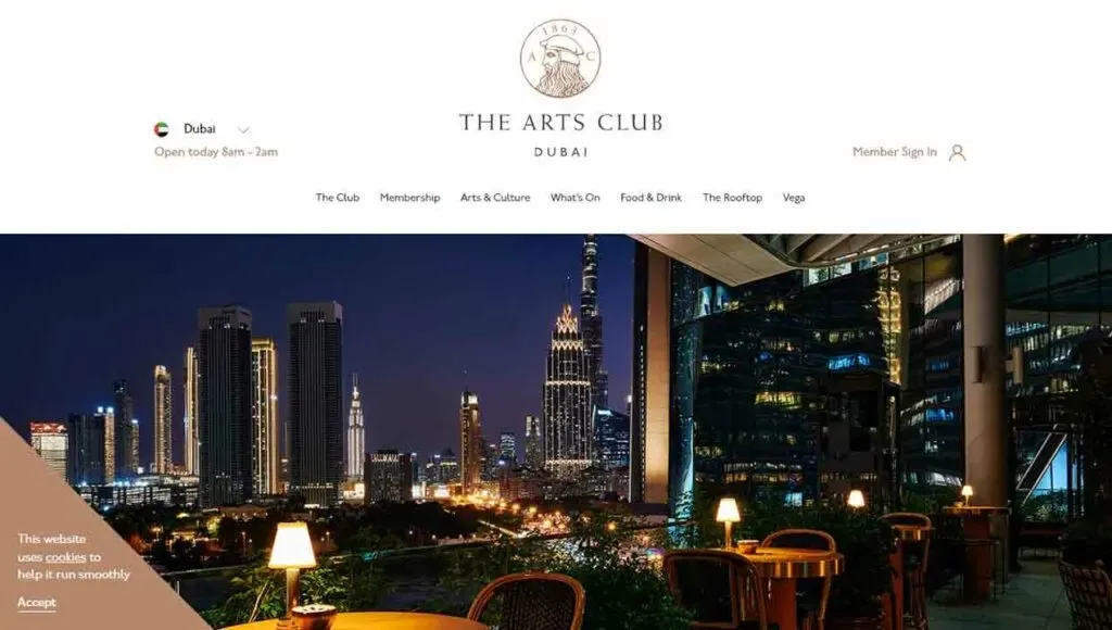 A nighttime view of Dubai's skyline features illuminated skyscrapers, including the distinctive Burj Khalifa, set against a clear sky, with greenery and seating visible in the foreground on a balcony of The Arts Club Dubai.