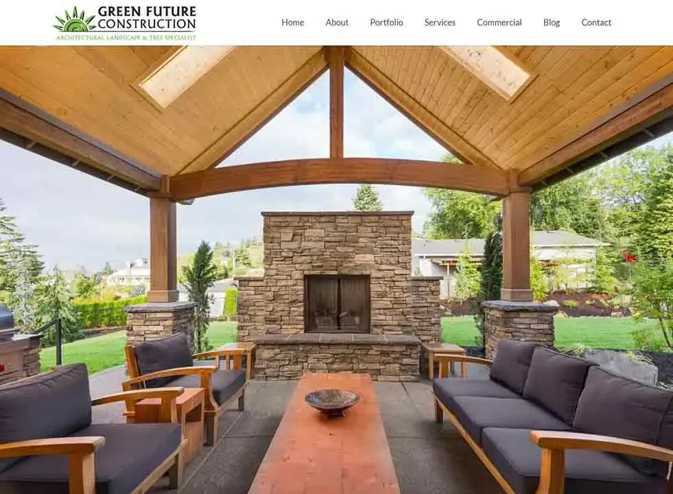 The image showcases a modern outdoor patio with a wooden cathedral ceiling, featuring a central stone fireplace flanked by lush green landscaping, and surrounded by comfortable wooden seating, under a clear sky.