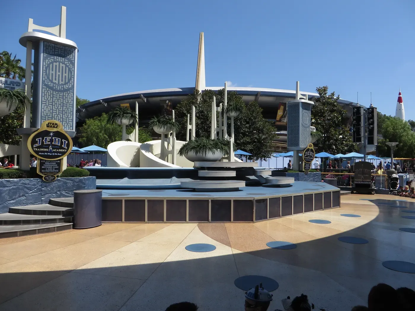 Área de escenario al aire libre en un parque temático con arquitectura futurista y señalización, enmarcado por un cielo azul claro, con patrones circulares en el suelo y rodeado de vegetación y árboles.