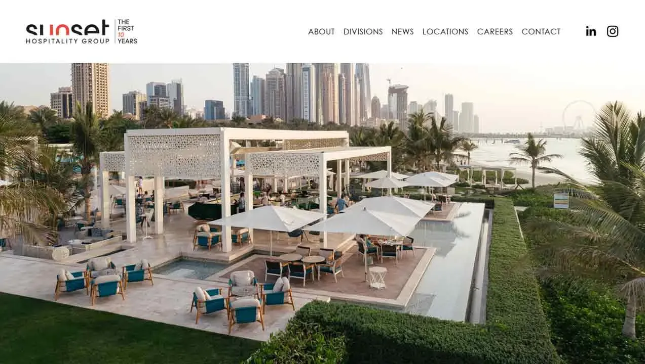 An elegant outdoor lounge area with modern white cabanas, plush seating, and umbrellas overlooks a lush green landscape and a distant city skyline.