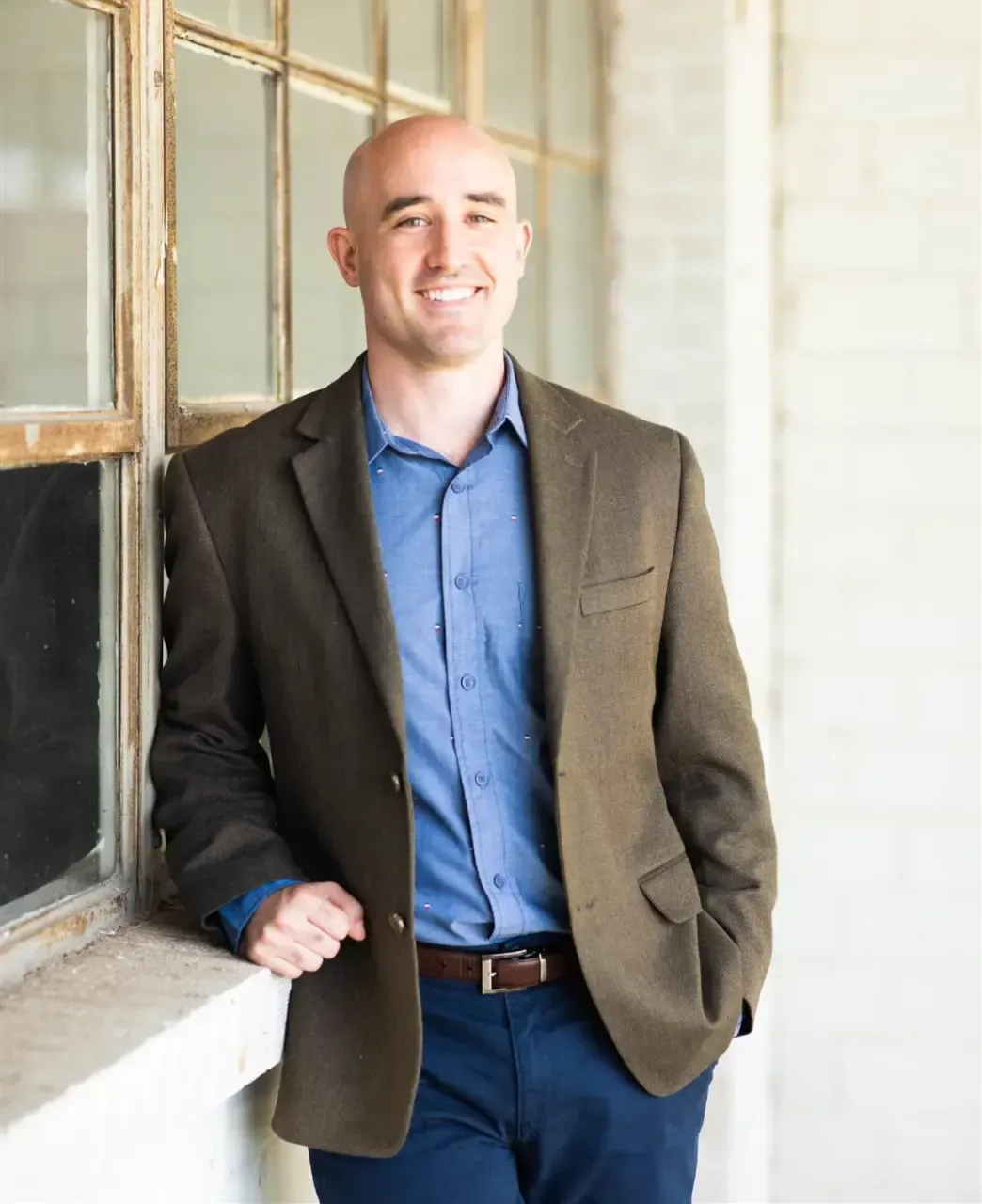 Un hombre sonriente con un blazer marrón y camisa azul se encuentra casualmente junto a una gran ventana en un entorno industrial moderno y luminoso.