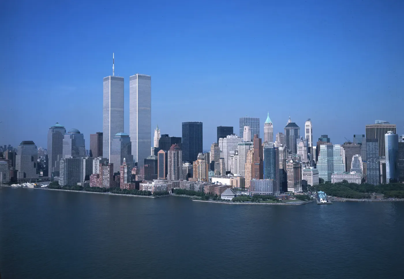 A panoramic view of the New York City skyline features the iconic Twin Towers of the World Trade Center, set against a clear blue sky with the calm waters of the Hudson River in the foreground.