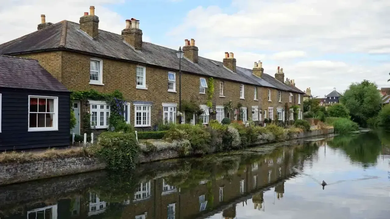 Una pintoresca fila de casas adosadas británicas tradicionales con fachadas de ladrillo bordean un tranquilo canal adornado con vegetación, bajo un cielo parcialmente nublado, reflejando el encanto de la arquitectura rural y la vida serena junto al agua.