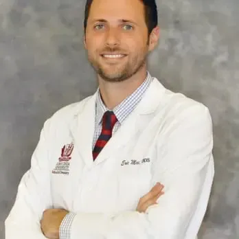 Un dentista masculino sonriente con una bata blanca con el logo de una universidad se muestra confiado con los brazos cruzados contra un fondo gris y texturizado, vistiendo una camisa a cuadros y una corbata a rayas.