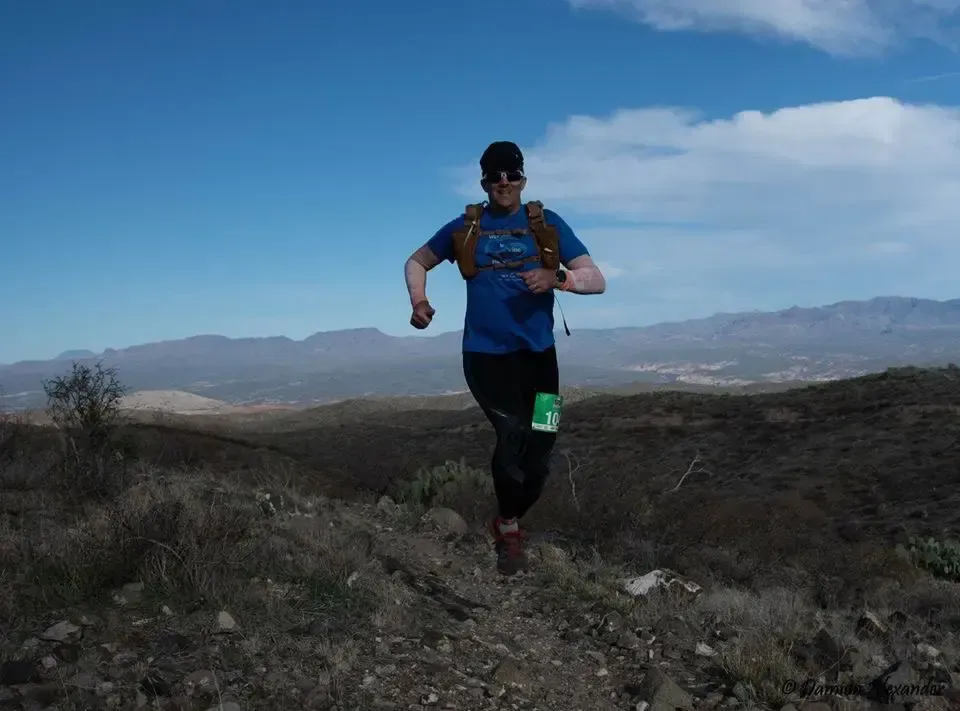 Un corredor con una camiseta azul y un paquete de hidratación corre por un sendero rocoso a través de un paisaje árido y montañoso bajo un cielo azul claro, encarnando el espíritu de la aventura al aire libre y el fitness.