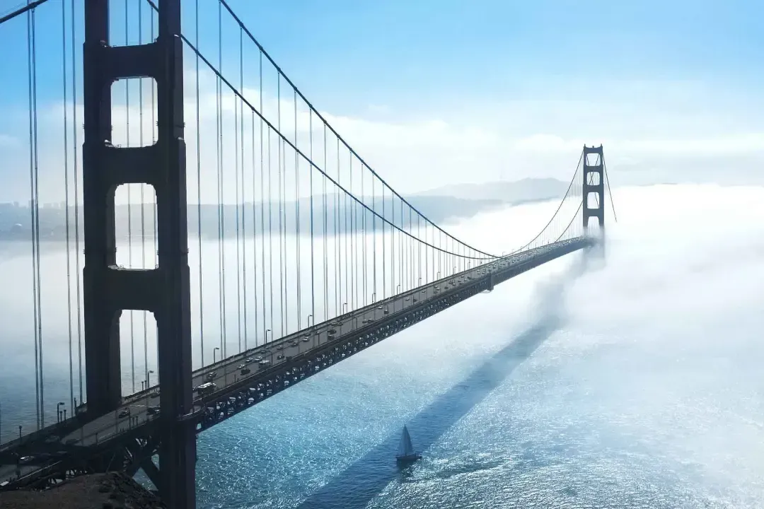 Un majestuoso puente colgante se extiende sobre una bahía iluminada por el sol, parcialmente envuelto en niebla, con un solitario velero navegando sus tranquilas aguas abajo.