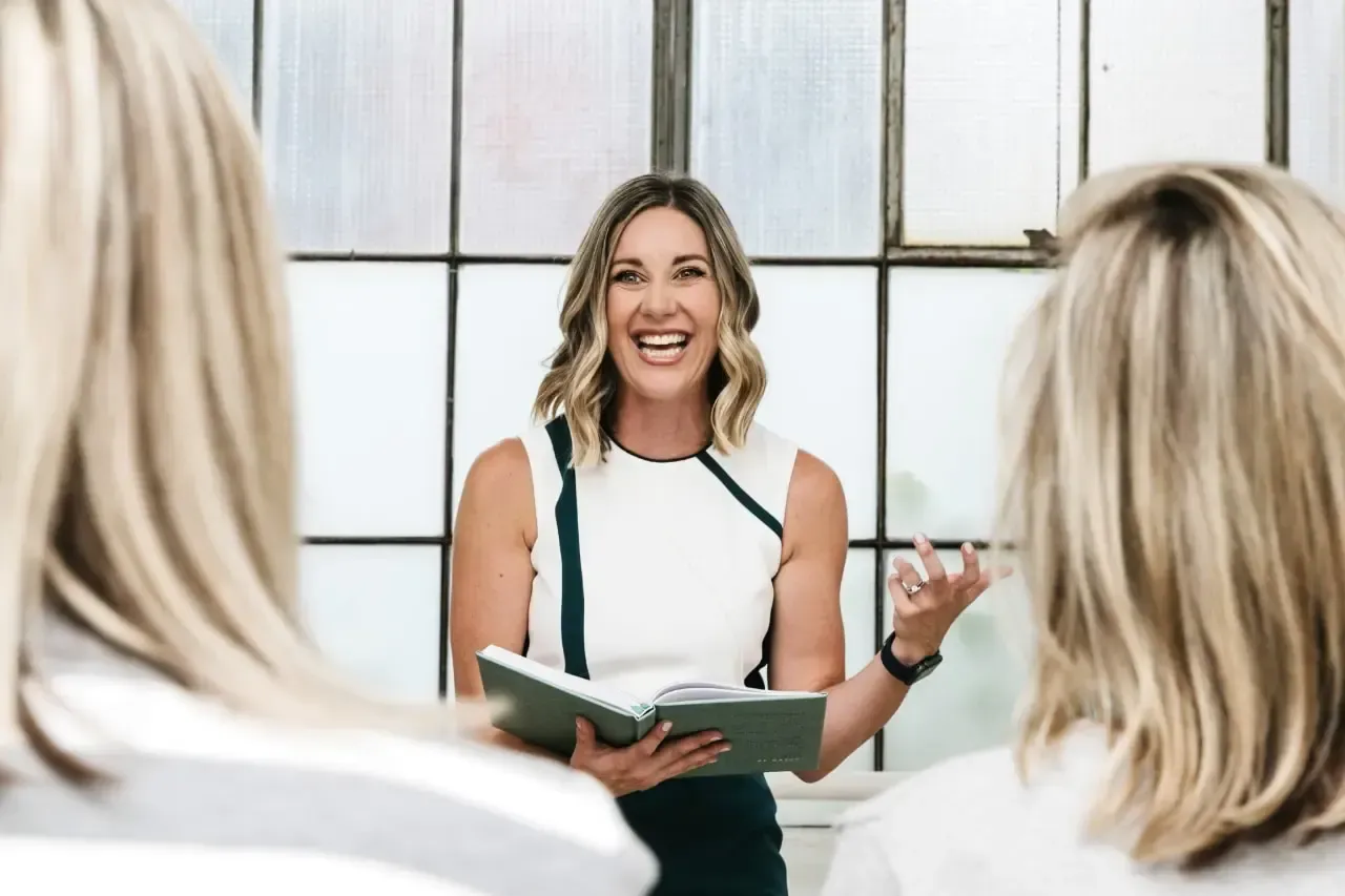 A woman standing in a bright room is energetically giving a speech while holding an open book, with two people listening in the foreground.