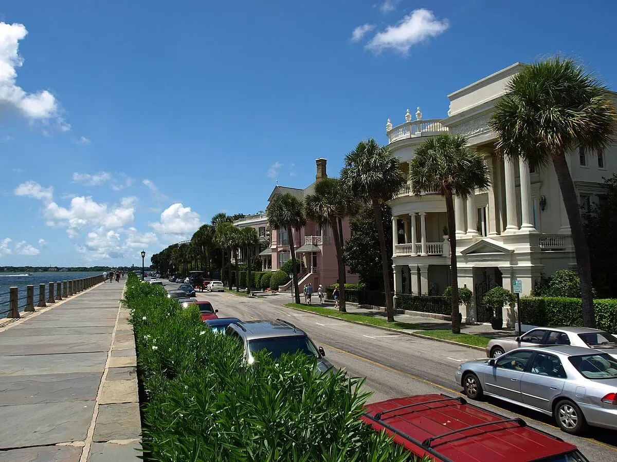 Casas históricas de estilo colonial bordean una pintoresca calle en Charleston, Carolina del Sur, con vista al paseo marítimo y palmeras bajo un cielo azul brillante.