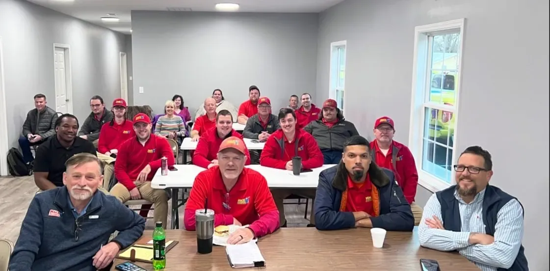 Un grupo diverso de personas con uniformes rojos se sientan atentamente en mesas en una sala de conferencias luminosa, participando en una reunión.