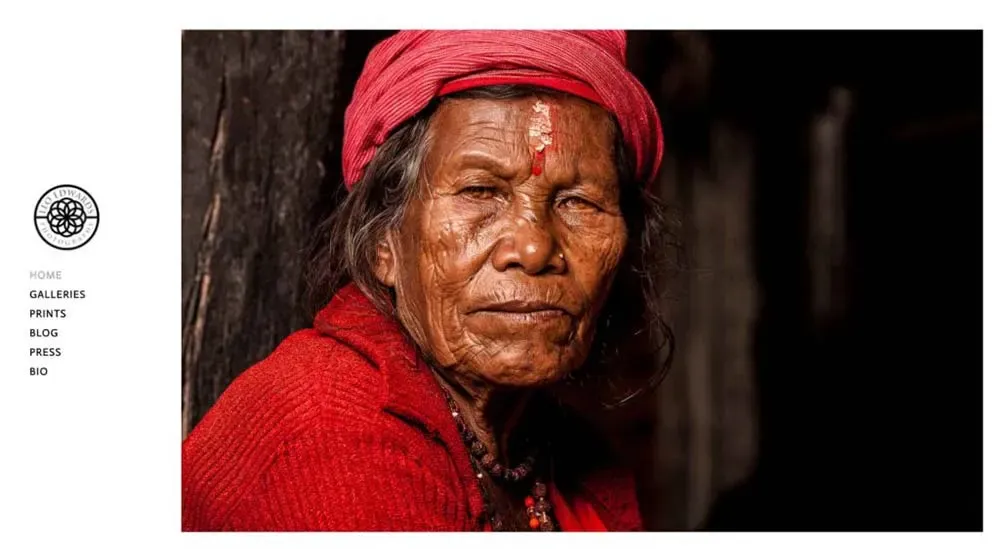 An elderly person wearing a red hat and sweater gazes contemplatively, with a dark background highlighting their weathered face and rich cultural attire.