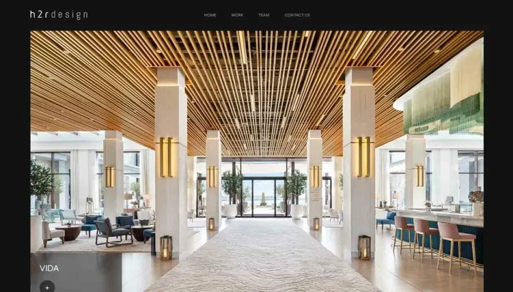 A spacious, modern hotel lobby features a wooden slatted ceiling, elegant pendant lighting, tall white pillars, and an open layout with plush seating areas, leading to large glass doors and scenic views beyond.