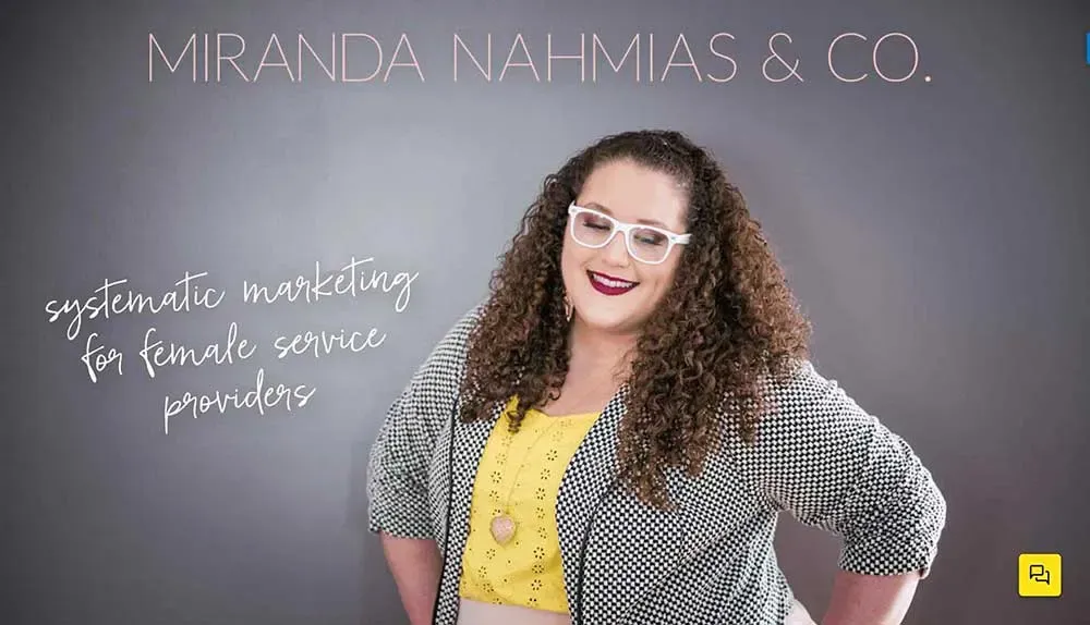A woman with curly hair, wearing glasses, a yellow top, and a black-and-white patterned jacket, stands confidently with hands on hips against a gray backdrop featuring the text "Miranda Nahmias & Co: Systematic Marketing for Female Service Providers."