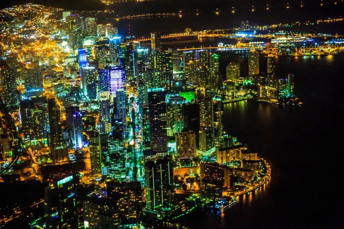 Esta vibrante vista aérea captura un deslumbrante paisaje urbano por la noche, con rascacielos iluminados por luces coloridas que se reflejan en el frente marítimo, mostrando la bulliciosa vida urbana y energía.
