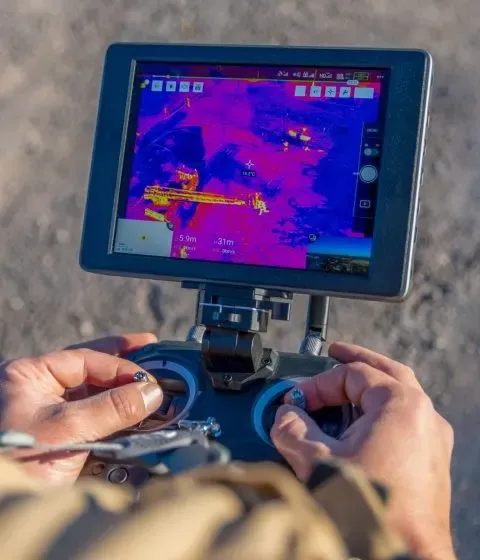 Una persona opera un controlador de dron que presenta una pantalla montada mostrando una vista vívida de imágenes térmicas de un paisaje, destacando variaciones de calor en colores brillantes contra un fondo neutro.