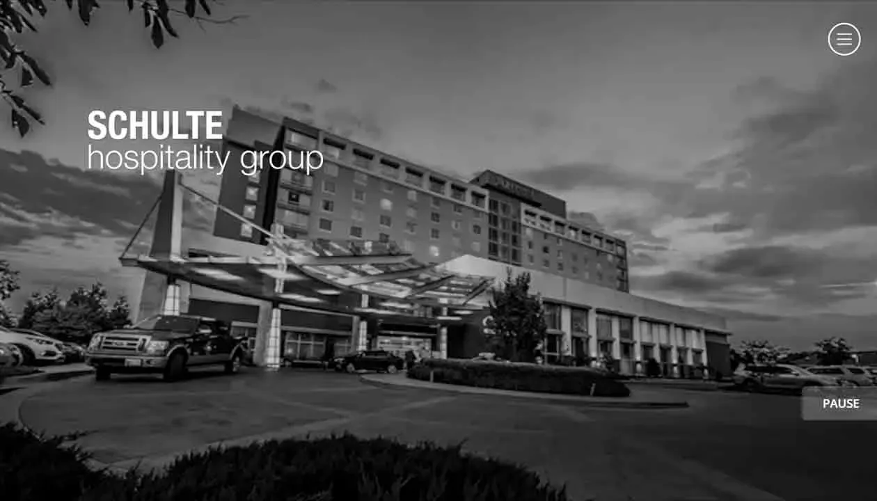 A black and white photo showcases the exterior view of a large hotel building with a modern glass canopy entrance, surrounded by parked cars and landscaped greenery, under a partly cloudy sky.