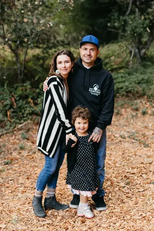 Una familia de tres posa junta en un entorno forestal, rodeada de exuberante vegetación y un manto de hojas caídas, con los padres parados cerca de su hijo pequeño que sonríe a la cámara.