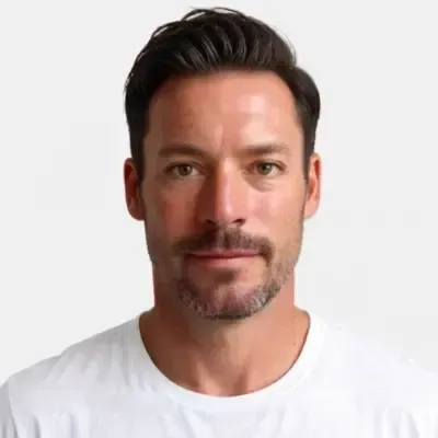 A man with short dark hair and a beard is wearing a plain white T-shirt against a neutral gray background.
