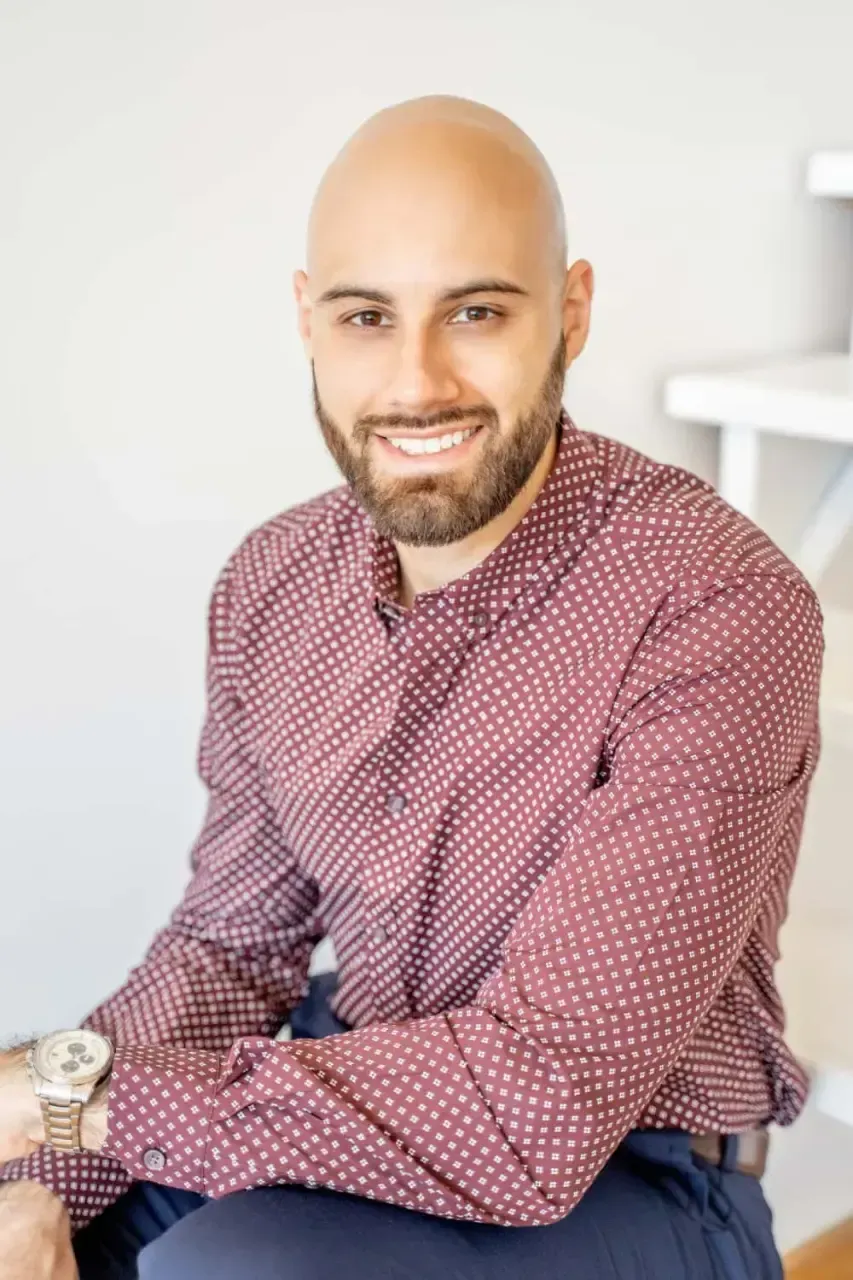 Un hombre calvo con barba y una sonrisa alegre está sentado contra un fondo claro, vistiendo una camisa marrón con estampado y pantalones azules, irradiando un comportamiento profesional y accesible.