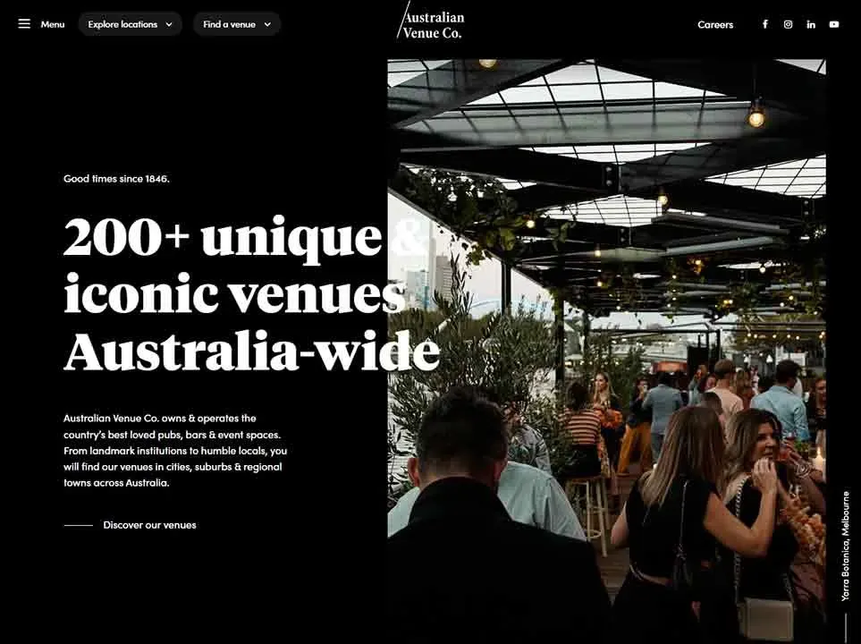 A bustling, indoor venue is filled with people socializing under a ceiling adorned with hanging plants and festoon lights, creating a lively and atmospheric setting for a celebratory event.