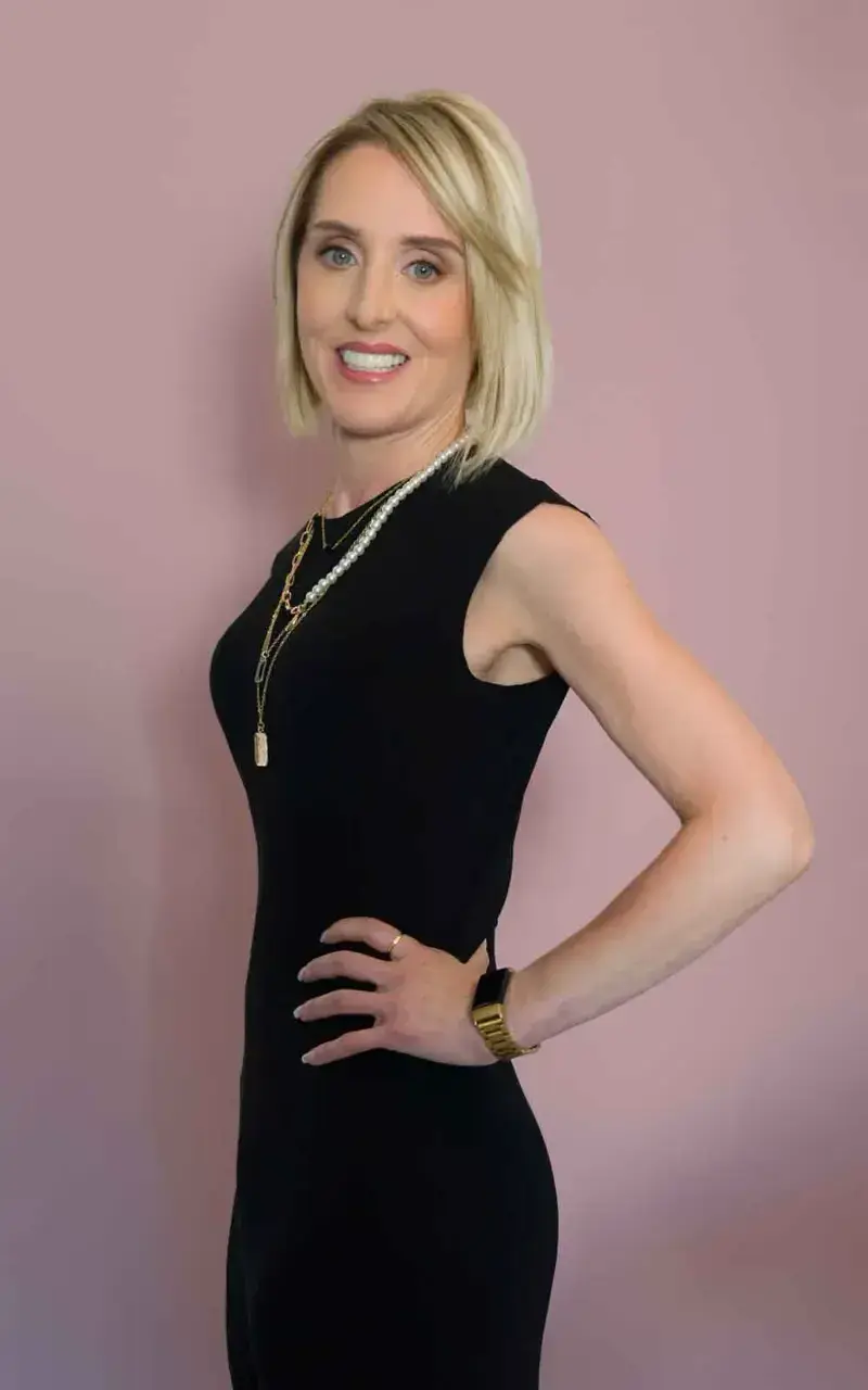Una persona con cabello rubio corto lleva un vestido negro sin mangas y collares en capas, de pie contra un fondo rosa suave.