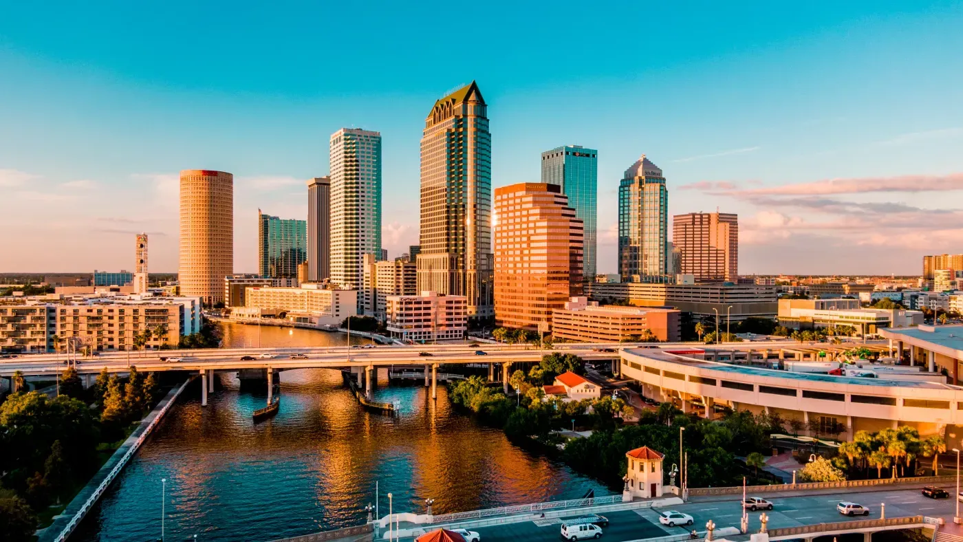 Un vibrante horizonte del centro de Tampa bajo un cielo azul claro, con modernos rascacielos reflejando la luz del sol, arquitectura urbana y un puente prominente cruzando un río sereno.
