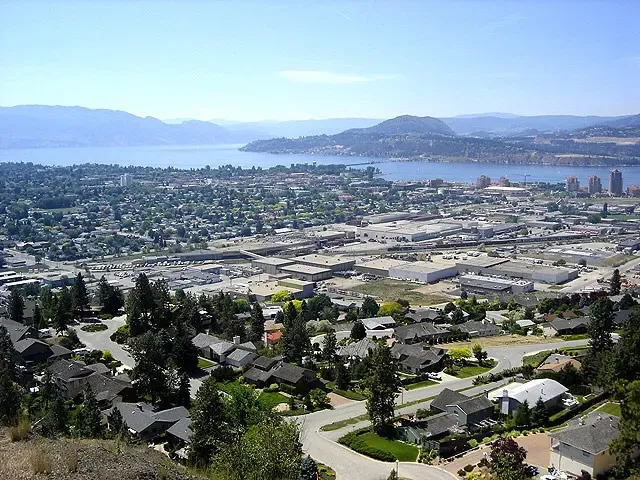 A scenic aerial view of Kelowna showcases a sprawling urban landscape nestled between lush greenery and expansive waters, with mountains in the background under a clear blue sky.