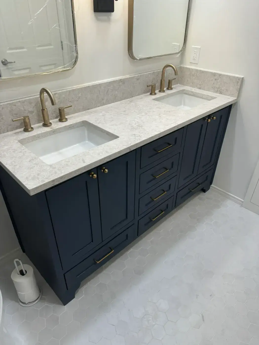 Un baño moderno presenta un elegante tocador doble con una encimera blanca y lavabos rectangulares dobles, acentuados por elegantes grifos dorados y enmarcados por gabinetes azul oscuro, sobre un suelo de baldosas hexagonales.