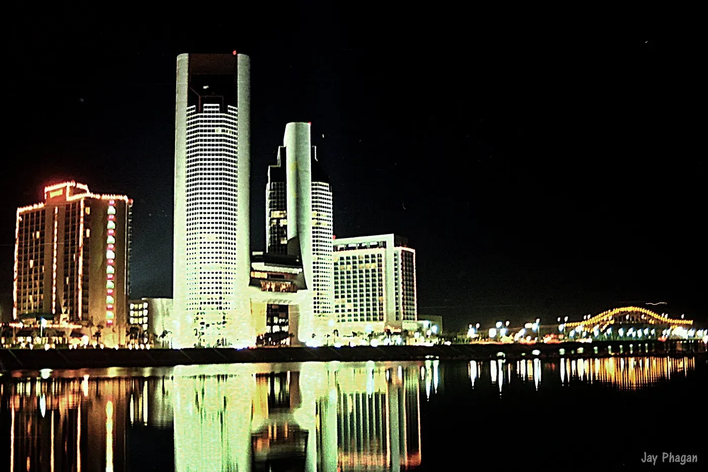 Vista nocturna del horizonte de Corpus Christi reflejándose en el agua, con modernos rascacielos iluminados y un puente distante iluminado, creando una escena de frente marítimo serena y pintoresca.