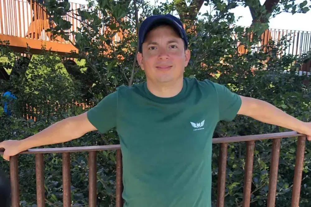 Una persona con una camisa y gorra verde está de pie al aire libre contra un fondo de vegetación exuberante y barandillas de madera, sonriendo a la cámara.
