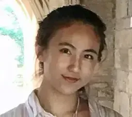 Una persona joven con cabello oscuro y largo, vistiendo una camisa de rayas de color claro, sonríe suavemente mientras está de pie frente a un arco de piedra con luz solar filtrándose, creando una atmósfera cálida y acogedora.