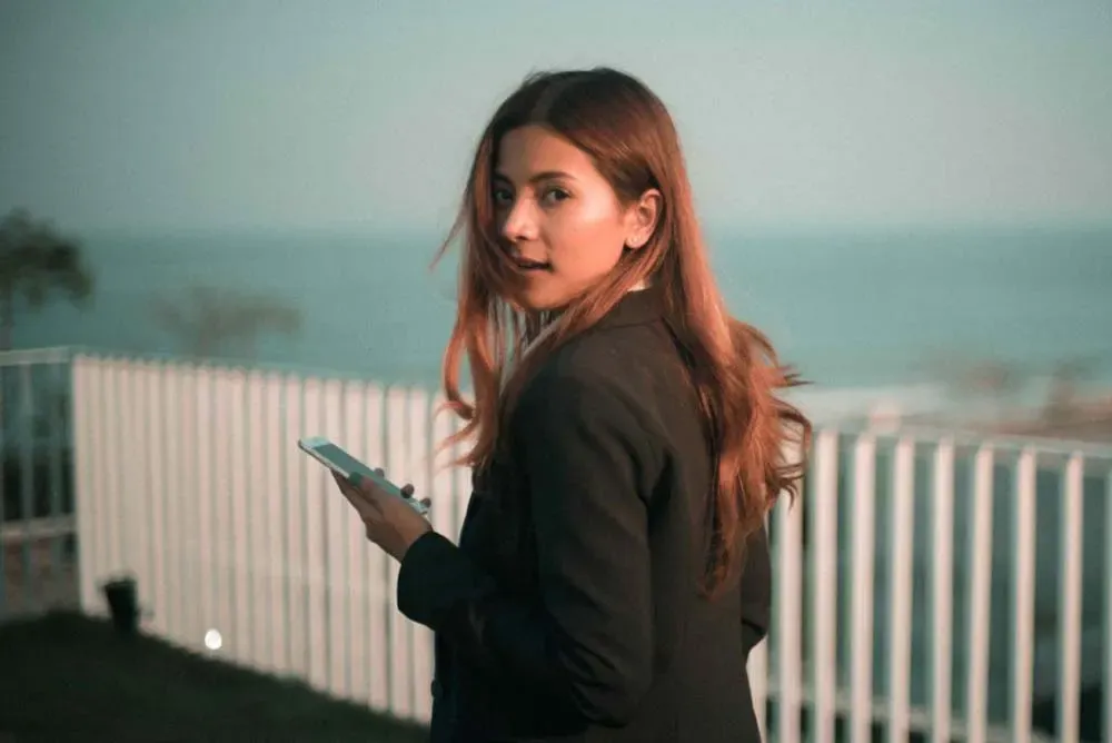 Mujer joven sosteniendo un teléfono mira por encima del hombro junto a una cerca frente al océano al anochecer