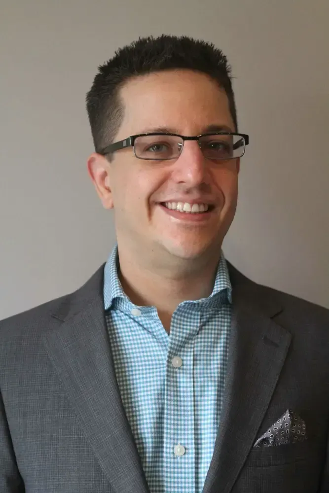 Una persona sonriente con gafas y un blazer gris sobre una camisa a cuadros azul se encuentra frente a un fondo liso.