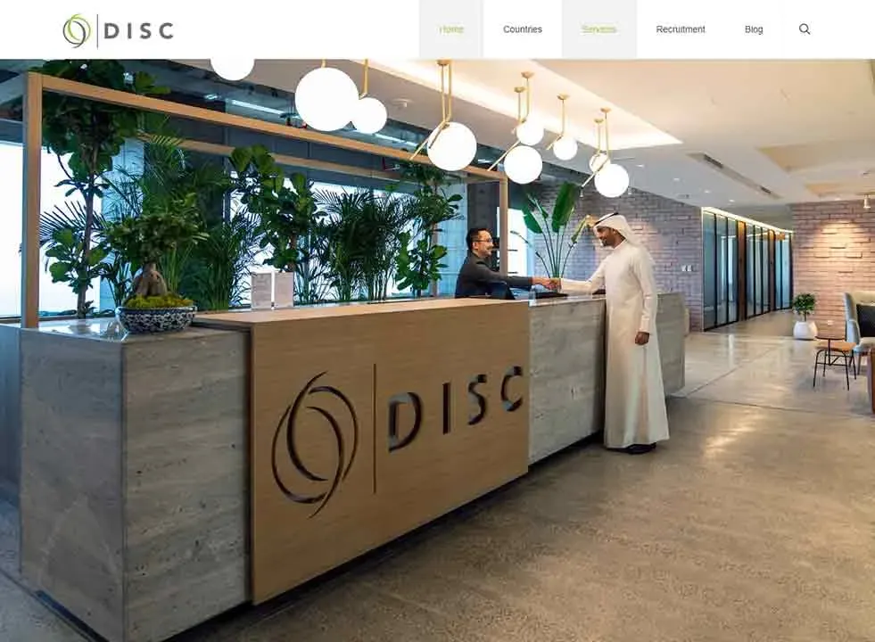 A modern office lobby with a large reception desk is adorned with green plants and spherical pendant lights, where a person in a dark outfit interacts with another wearing traditional attire, against a backdrop of stone accents and warm lighting.