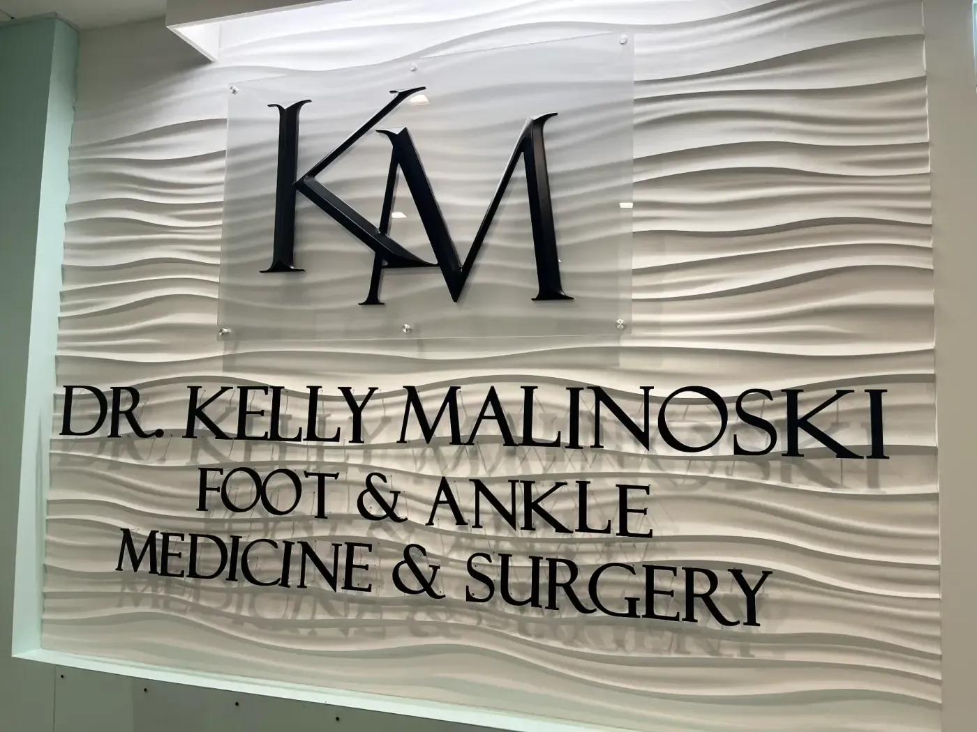 Un panel de pared elegante y moderno muestra las letras "KM" de manera prominente, con el texto "Dr. Kelly Malinoski Medicina y Cirugía del Pie y Tobillo" en letras negras y audaces contra un fondo blanco texturizado con patrón de ondas, enfatizando una práctica médica profesional especializada en podiatría.