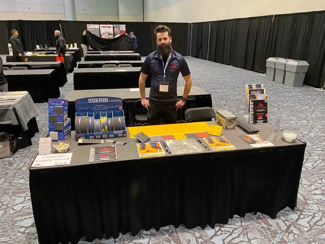 Un hombre con barba está detrás de una mesa de exhibición mostrando herramientas industriales y folletos informativos, en una gran sala de conferencias con cabinas cubiertas de negro.