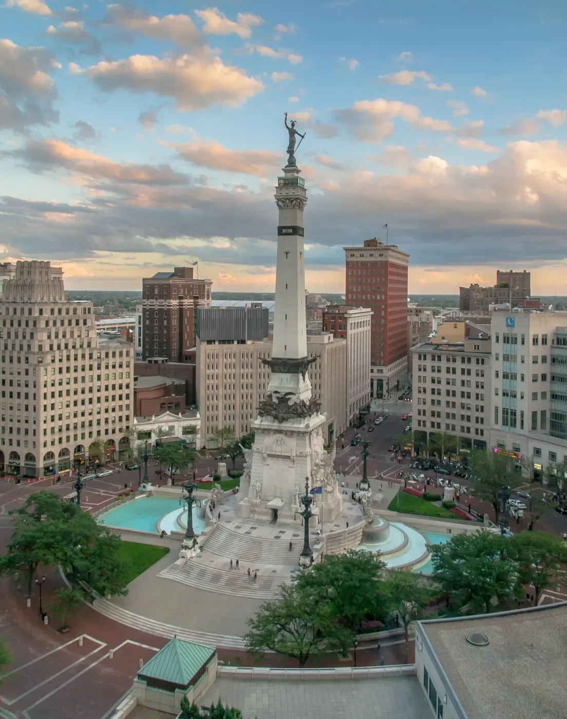 Vista aérea de Monument Circle en Indianápolis, mostrando el icónico Monumento a los Soldados y Marineros rodeado de edificios históricos y exuberante vegetación bajo un cielo parcialmente nublado.